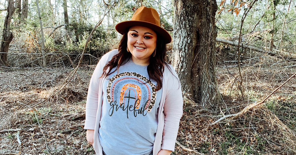 Capital One associate Danika stands in front of a tree wearing a hat and a shirt that says "grateful"